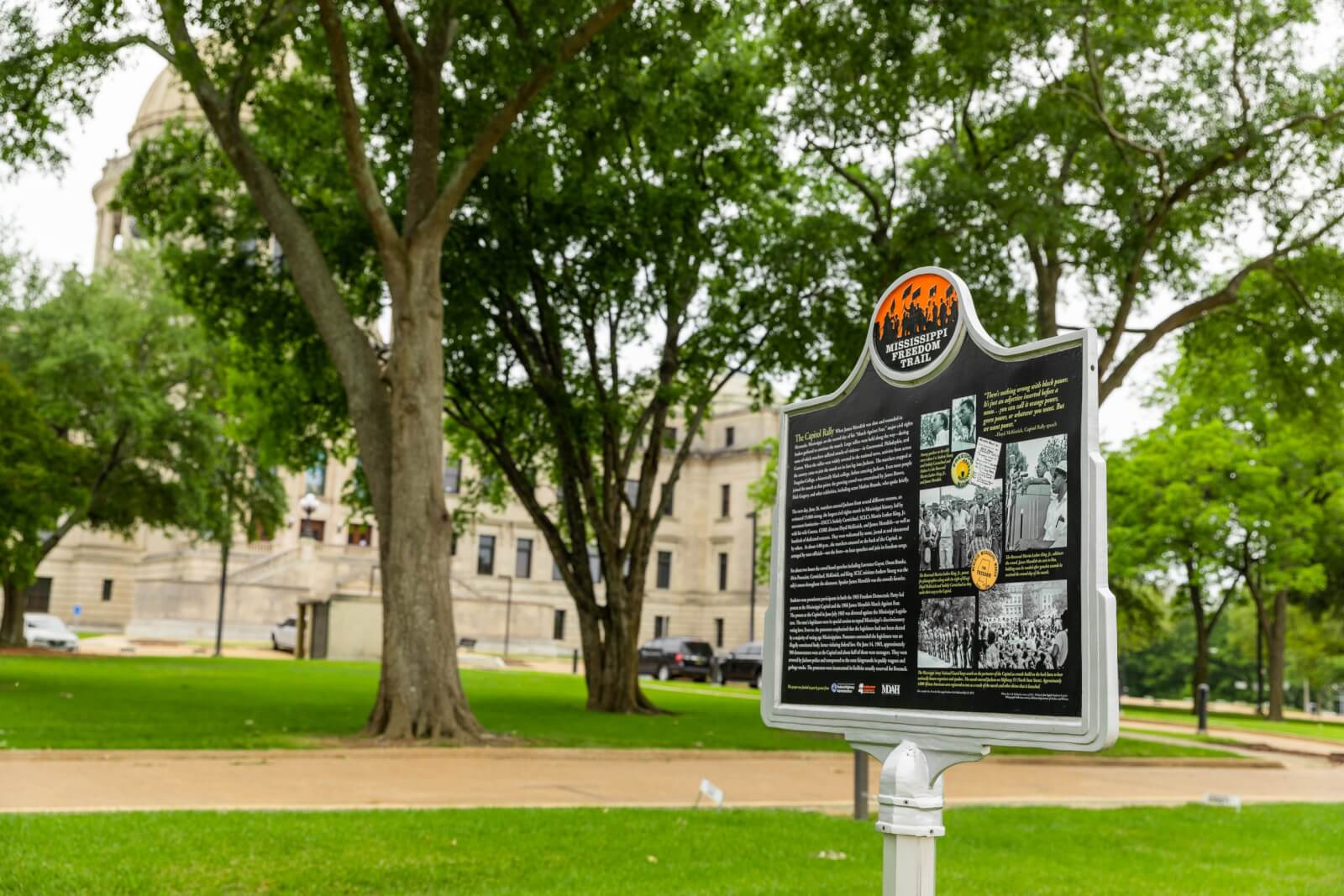 A historical marker sign from the Mississippi Freedom Trail stands on a grassy area, framed by trees with a large building visible in the background. Text and photos on the sign tell stories of courage and resilience.