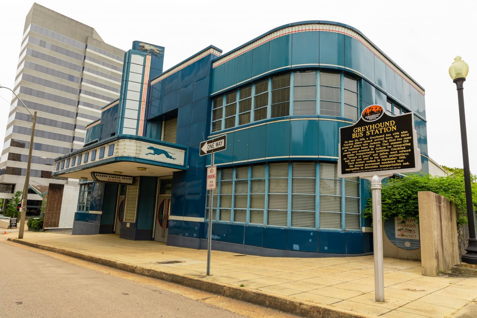 The historic Greyhound bus station, a site on the Mississippi Freedom Trail, features a blue Art Deco facade with curved windows and distinctive signage. It sits prominently on a city street, framed by a modern building in the background.