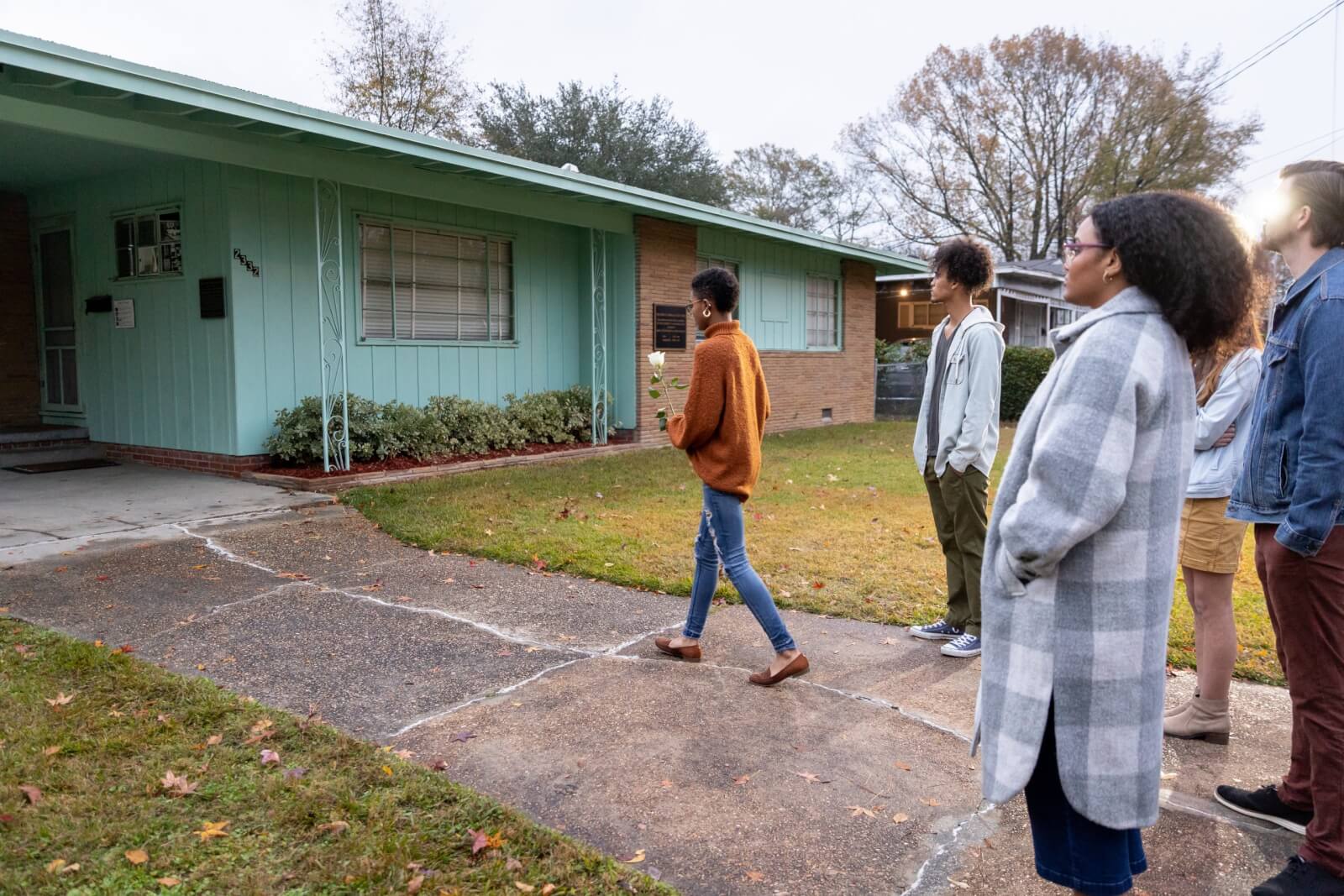A group of people stand outside a single-story teal-colored house, as if on a stop along the Mississippi Freedom Trail, with one person leading them up the path.