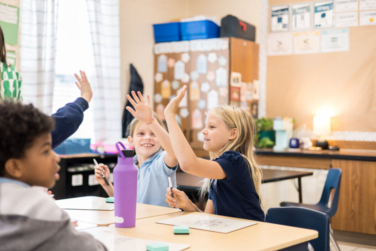 Nashville Private School Open House Dates - 2 Three children sit at a classroom table with whiteboards in a vibrant school setting, two smiling and raising their hands, while another looks on. Classroom supplies and educational posters are visible in the background.