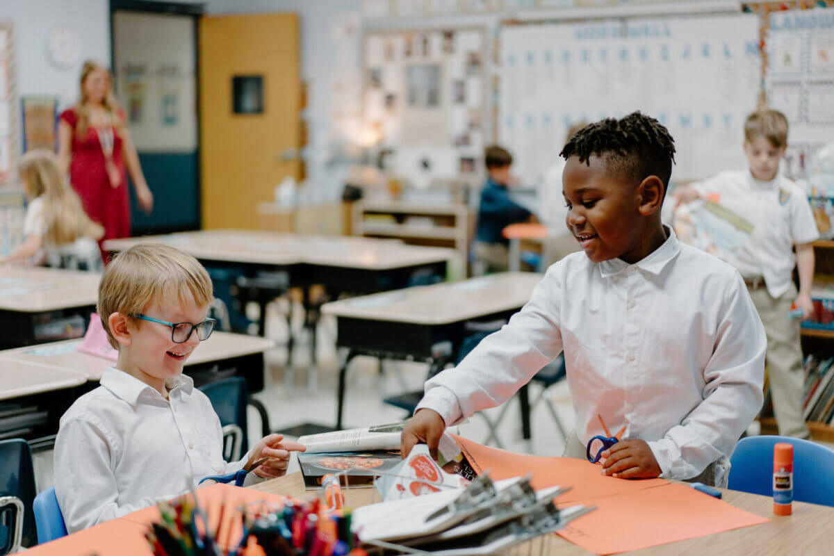 Nashville Private School Open House Dates - 14 Two boys in white shirts work on a paper craft project at a classroom table. One is handing paper to the other, and both are smiling. Other children and a teacher are in the background.