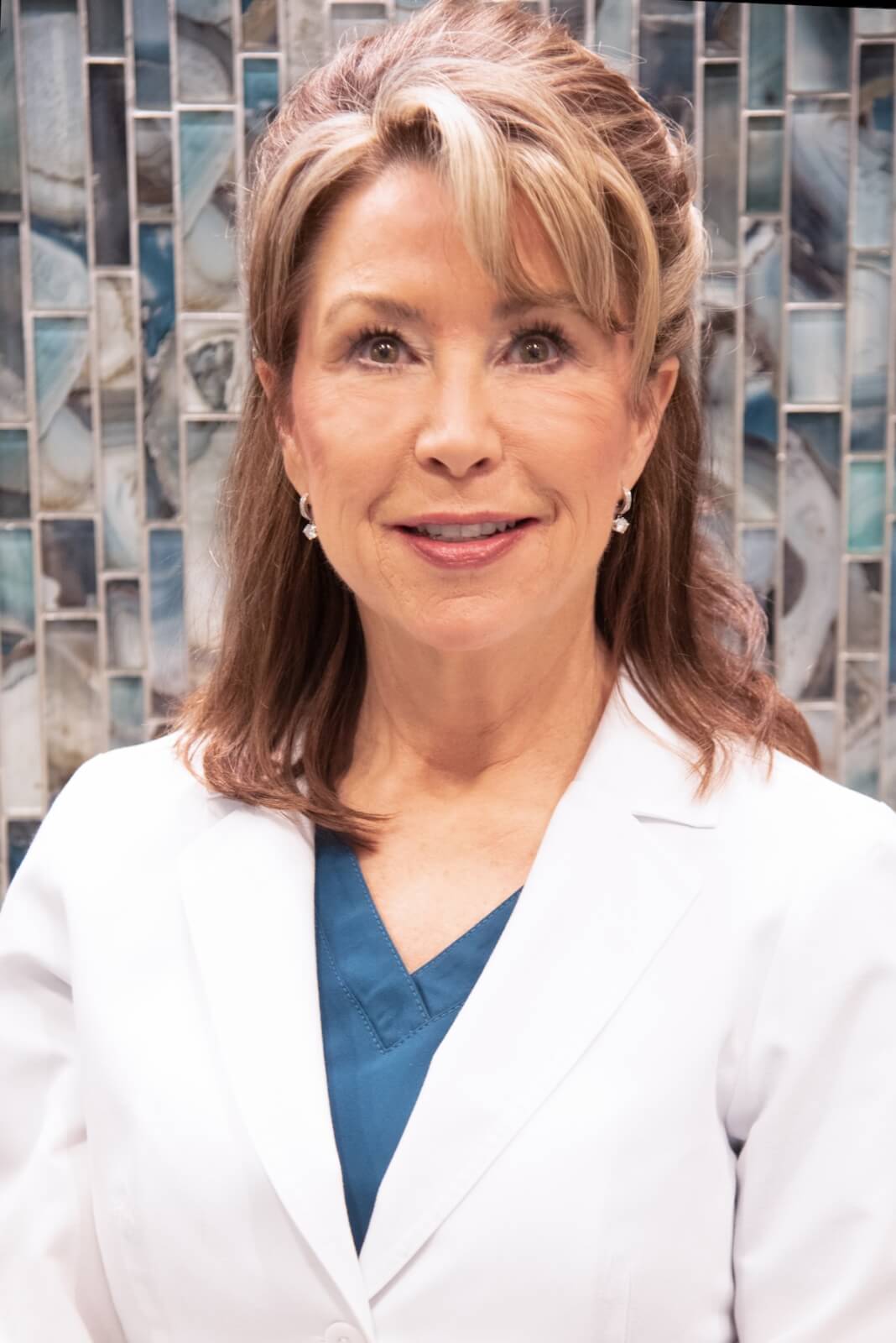 Kimberly Vaugh, President of HPIHair Partners, draped in a sleek white coat over a blue shirt, stands confidently before a patterned wall.