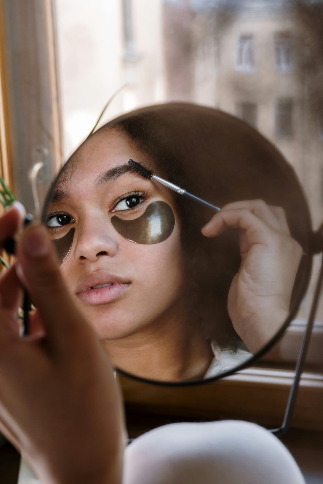 Applying makeup with a brush, she wears under-eye patches and looks into a round mirror by the window, reflecting on hair loss solutions.