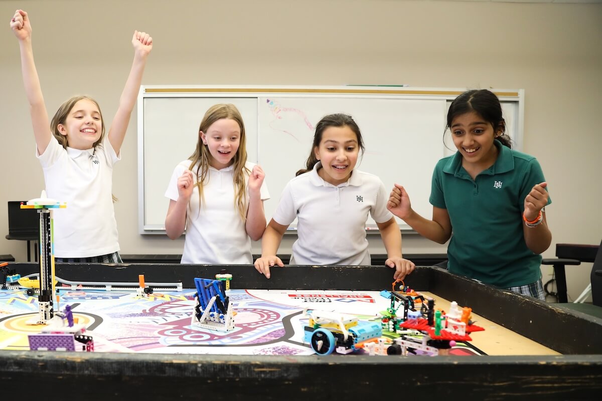 At Harpeth Hall, four girls cheer around a robotics competition table, where small robots are assembled and displayed. This vibrant scene perfectly embodies our vision statement that empowers women through hands-on learning and teamwork.