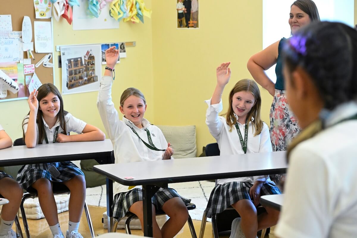 Students in uniforms sit at desks in a Harpeth Hall classroom, raising their hands. A teacher stands nearby, observing and empowering women to contribute confidently as part of the school's vision statement.