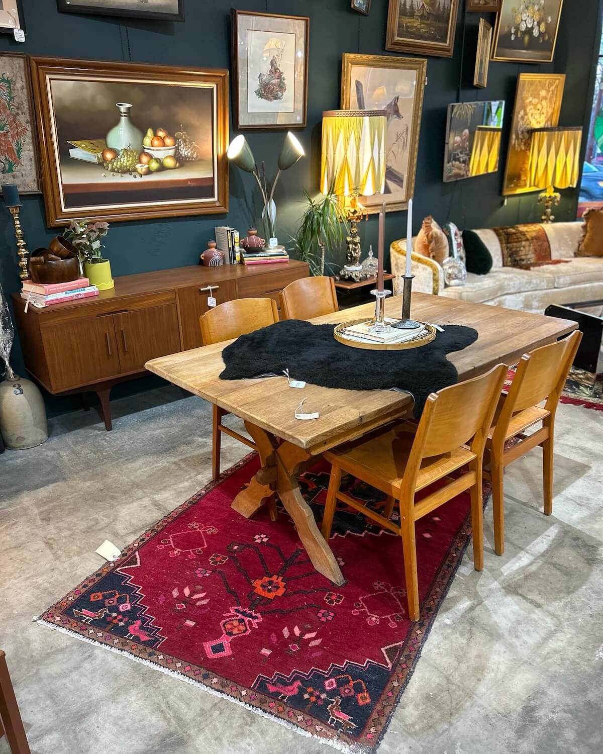 Nashville's Vintage Retail Trail: 8 Must-Visit Home Decor Spots - 1 A wooden dining table with six chairs rests on a red rug, enveloped by Nashville vintage furniture and framed art on dark walls. Two lamps with yellow shades grace a sideboard in the background.