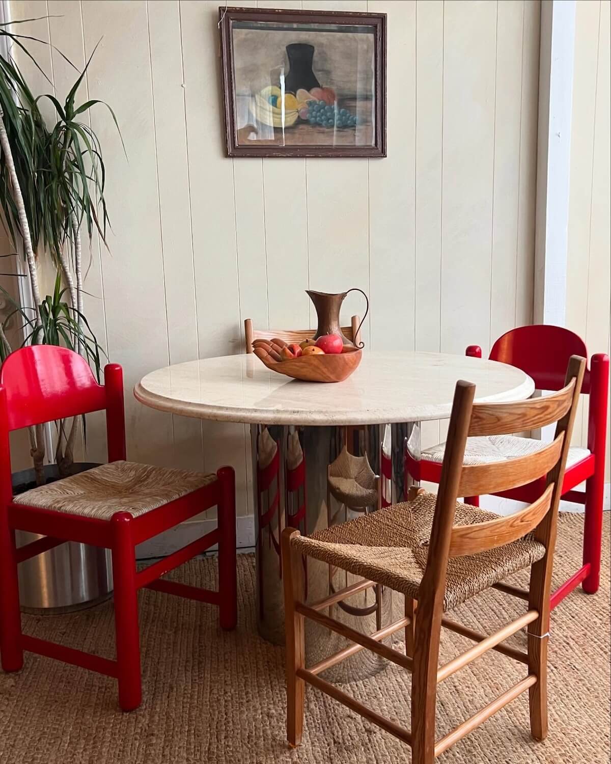 Nashville's Vintage Retail Trail: 8 Must-Visit Home Decor Spots - 2 A small round dining table with a reflective metal base, surrounded by three red chairs and a wooden one—a delightful find from Nashville vintage stores. A bowl of fruit and a metal jug sit elegantly on the table, beneath a painting hanging on the wall.