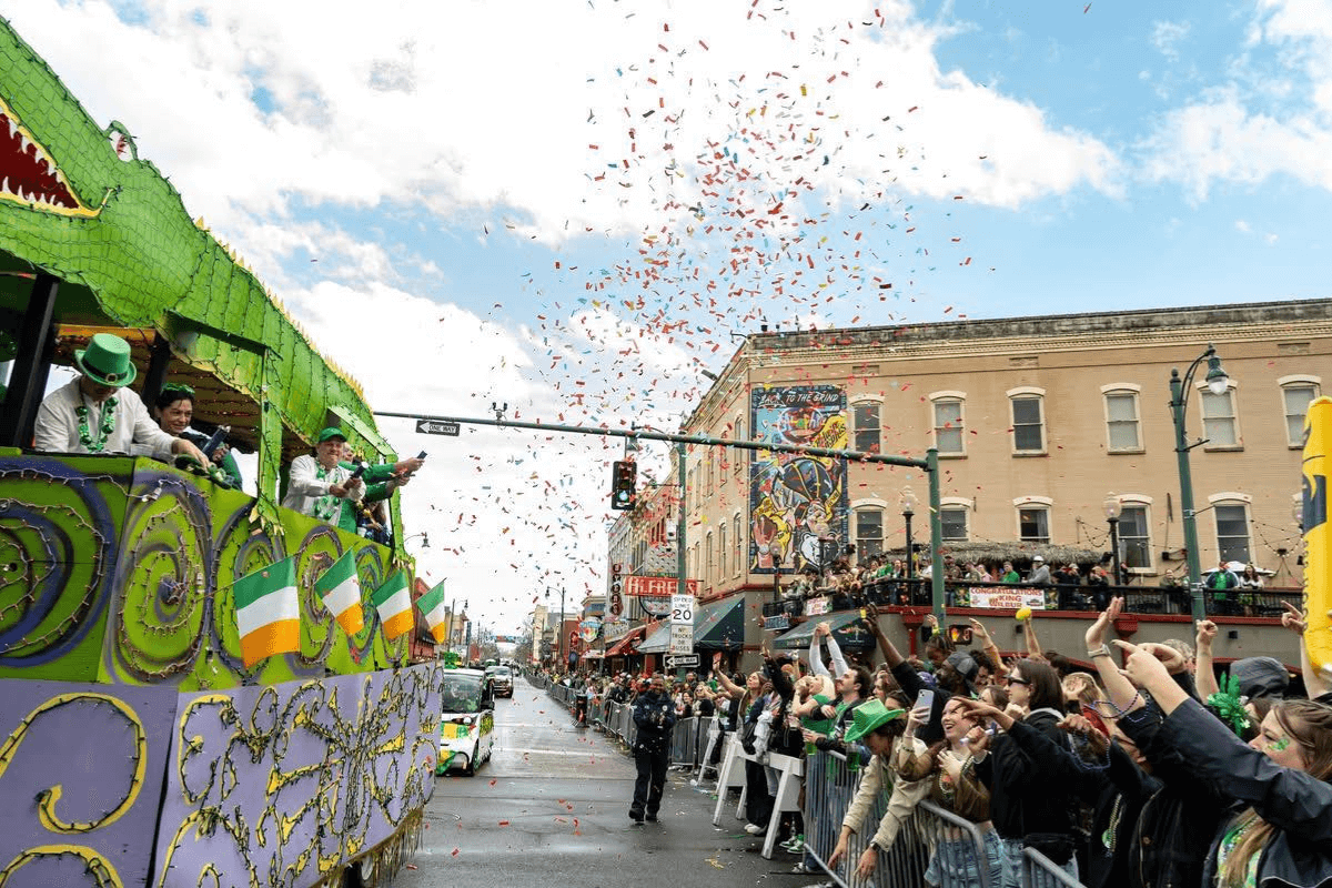 A parade float decorated in green and purple celebrates events in March, passing cheering crowds as confetti falls on a city street lined with brick buildings.