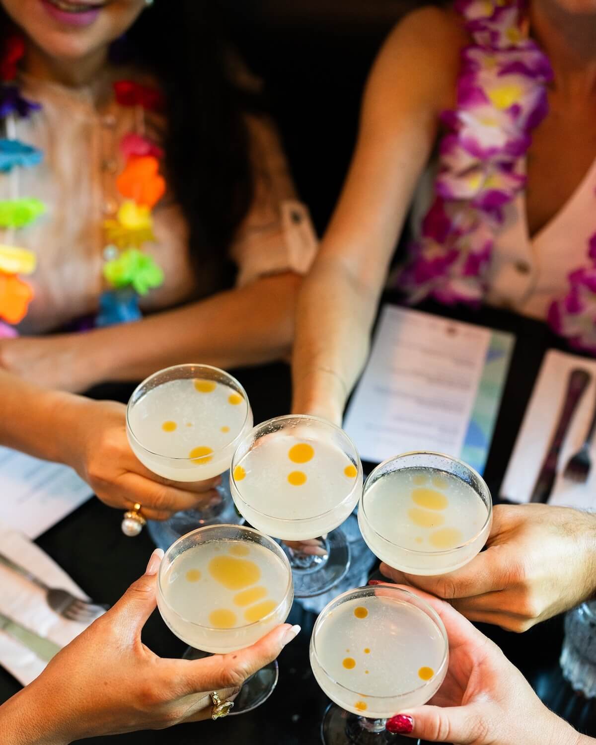 Five people holding glasses of a cocktail garnished with orange drops, surrounded by menus on a table, evoke the lively spirit of food and wine festivals.
