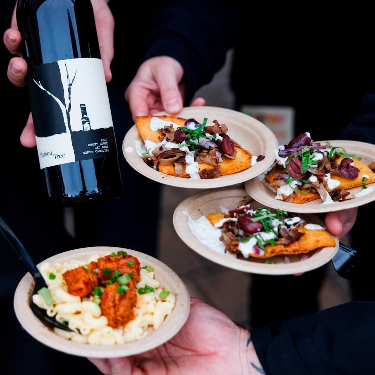 Four hands hold a bottle of red wine and three plates of food, reminiscent of a lively food and wine festival scene. The spread includes a pasta dish and two flatbreads with toppings, all served on paper plates.