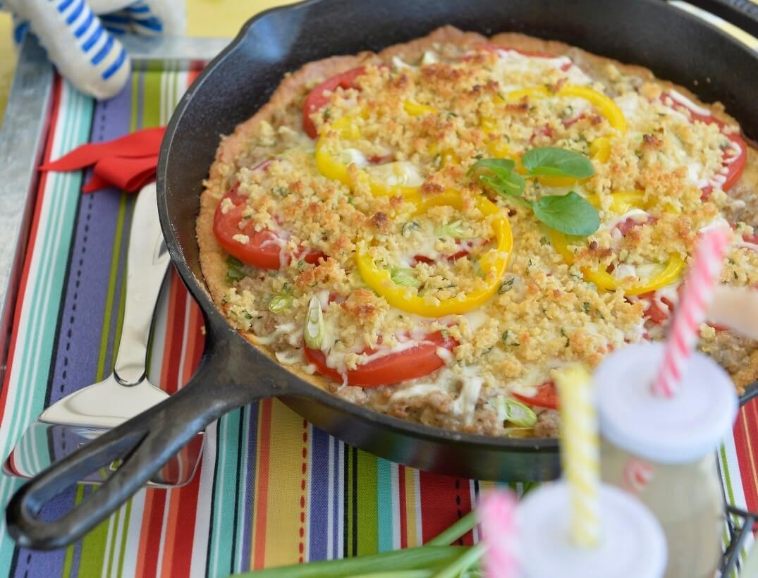 Cornbread in a skillet with tomato and yellow pepper slices, topped with cheese and a basil garnish, reminiscent of flavors celebrated at food and wine festivals, on a striped tablecloth.