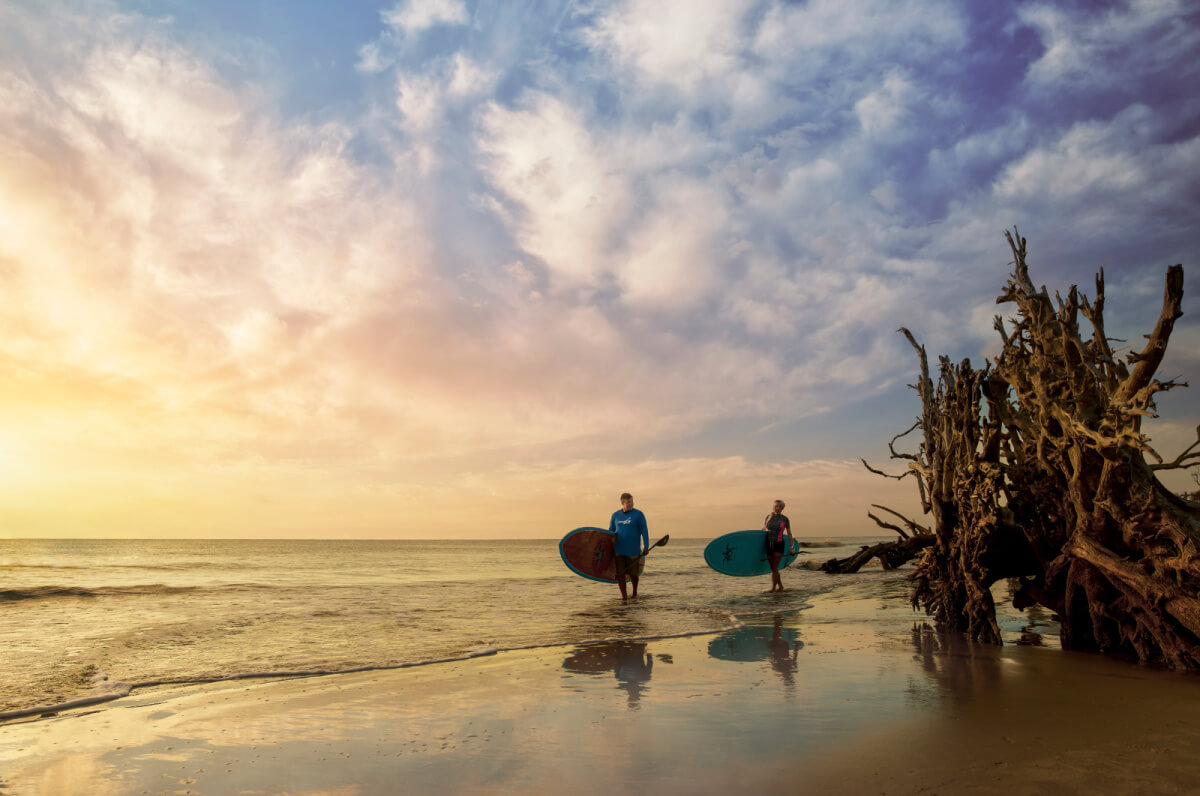 Two people stroll along a beach at sunset, surfboards in hand. A large driftwood tree stands on the right as the sky, painted with clouds and warm colors, mirrors the tranquil beauty often found along Southern waterways.