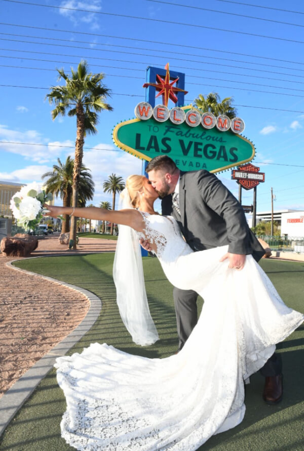They Had the Last Pre-COVID Wedding in Vegas! - 9 A couple in wedding attire poses in front of the "Welcome to Fabulous Las Vegas" sign, capturing the quintessential Las Vegas wedding moment. The groom dips the bride, who holds a vibrant bouquet, against a backdrop of palm trees and a clear sky.