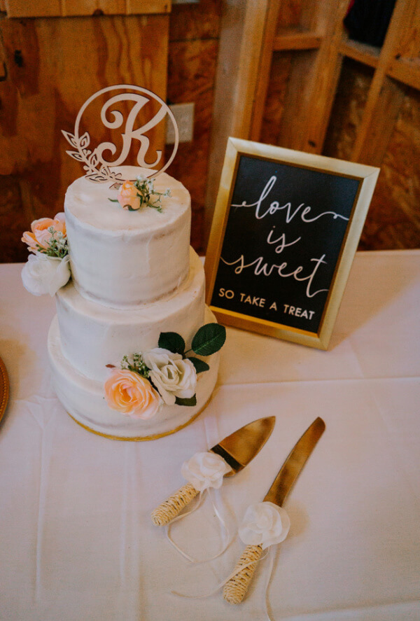They Had the Last Pre-COVID Wedding in Vegas! - 14 A three-tier white cake adorned with floral decorations and a letter "K" topper. Nearby, a framed sign reads "love is sweet so take a treat," accompanied by an elegant cake knife and server.