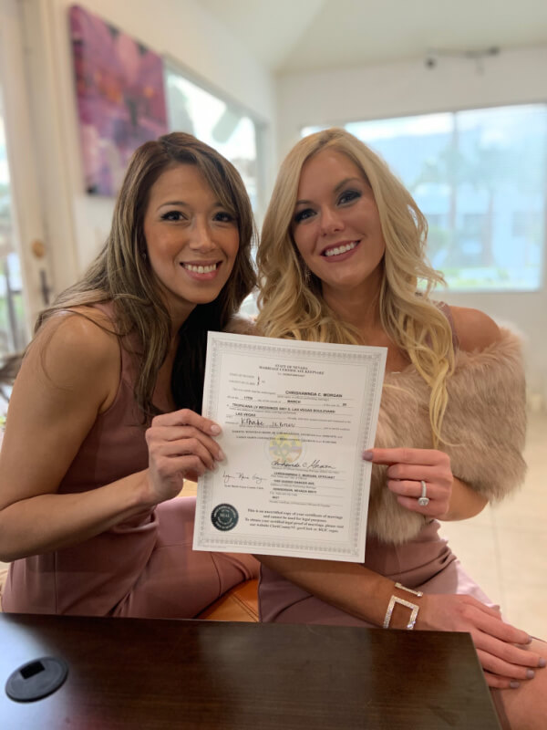 They Had the Last Pre-COVID Wedding in Vegas! - 5 Two women, elegantly dressed, are sitting indoors with radiant smiles as they hold a signed certificate celebrating a Las Vegas wedding.