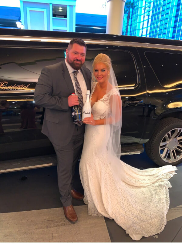 They Had the Last Pre-COVID Wedding in Vegas! - 8 Bride and groom stand in front of a black limousine, each holding a bottle. The bride dazzles in her white gown and veil, while the groom sports a gray suit with a patterned tie. It's the quintessential Las Vegas wedding, capturing the charm and spontaneity of love in Sin City.
