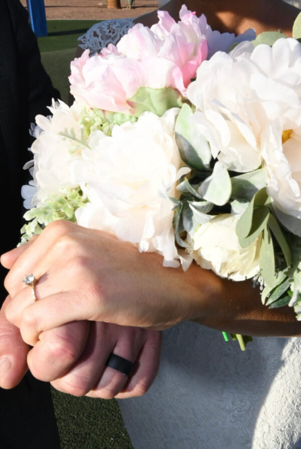 They Had the Last Pre-COVID Wedding in Vegas! - 6 A couple holding hands, one wearing a ring, stands against the backdrop of a beautiful Las Vegas wedding, complemented by a bouquet of white and pink flowers.