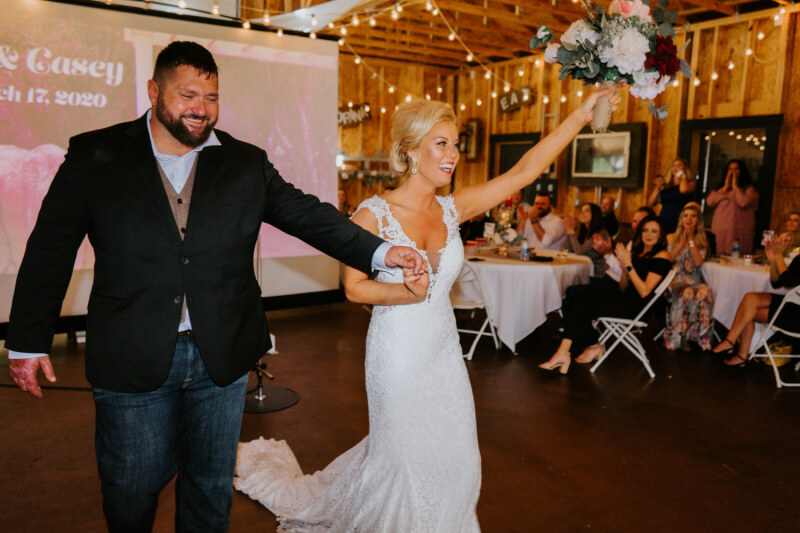 They Had the Last Pre-COVID Wedding in Vegas! - 15 Bride and groom hold hands and smile as they enter a warmly lit wedding reception venue, with guests seated at round tables applauding.