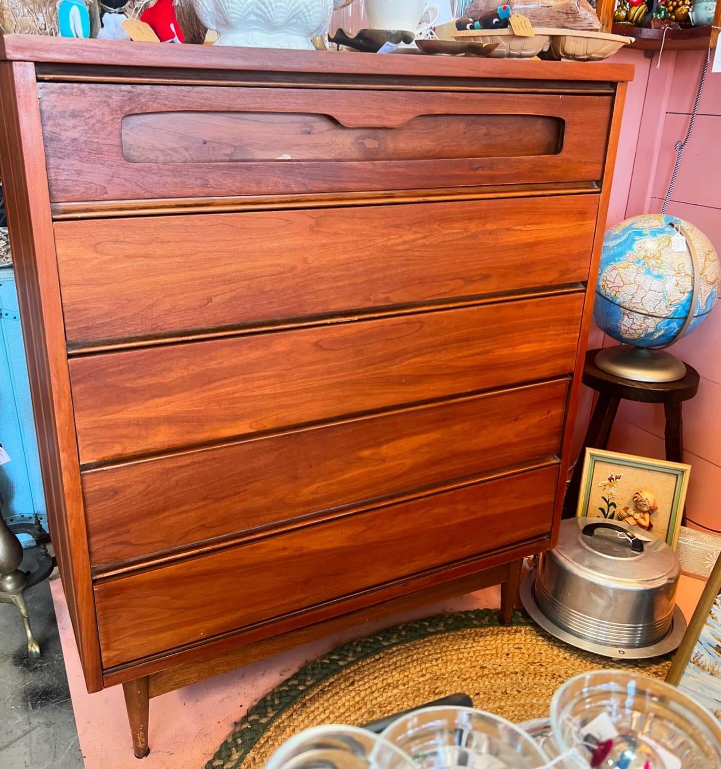 Nashville's Vintage Retail Trail: 8 Must-Visit Home Decor Spots - 7 A wooden chest of drawers from Nashville vintage stores stands in a room adorned with a globe, framed picture, and kitchenware on top. Nearby, stacked items like a cake tin and various knick-knacks add charm to this Nashville vintage furniture piece.