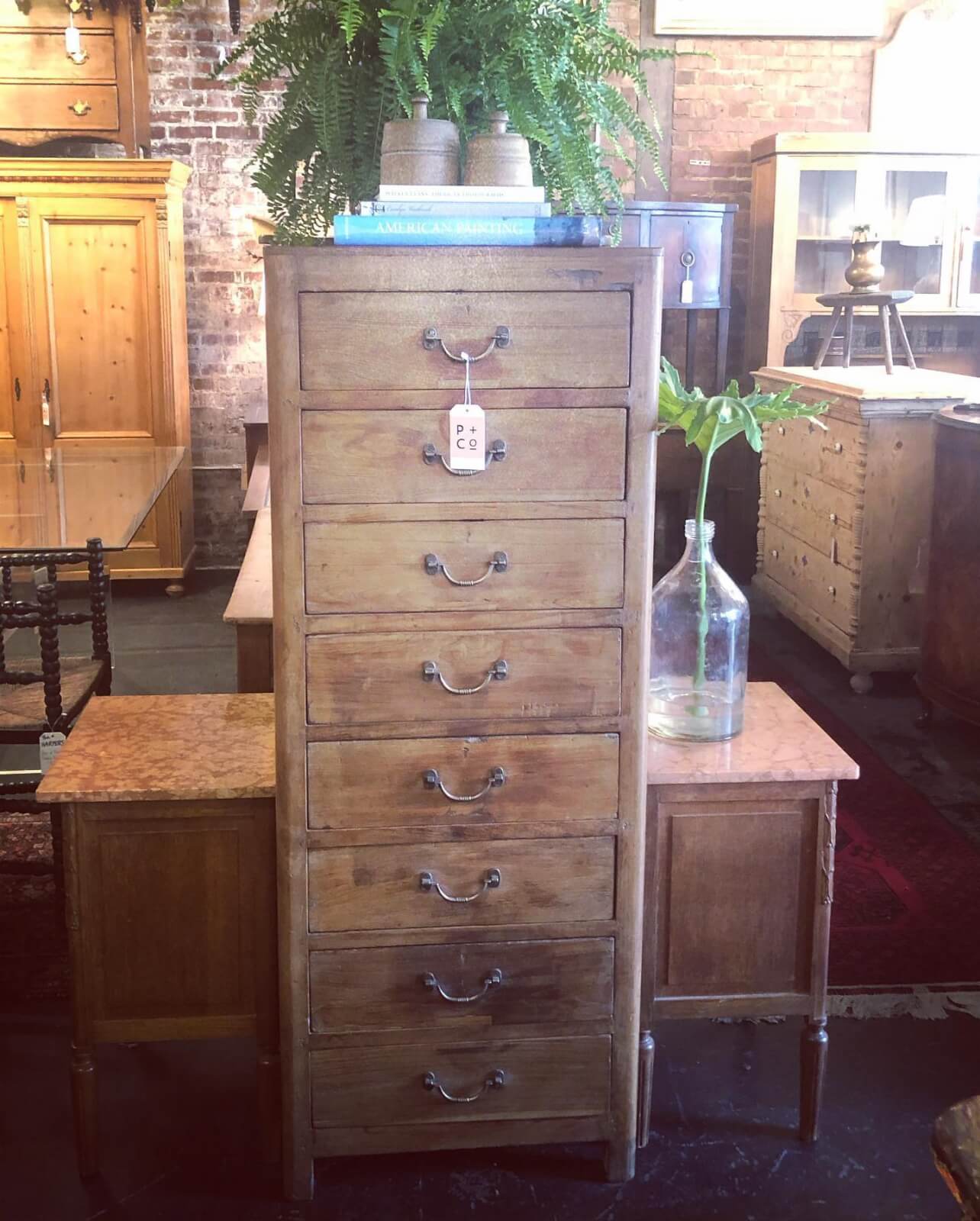 Nashville's Vintage Retail Trail: 8 Must-Visit Home Decor Spots - 4 A tall, narrow wooden chest of drawers with metal handles, topped with books and plants, is flanked by two smaller wooden tables in a Nashville vintage furniture store.