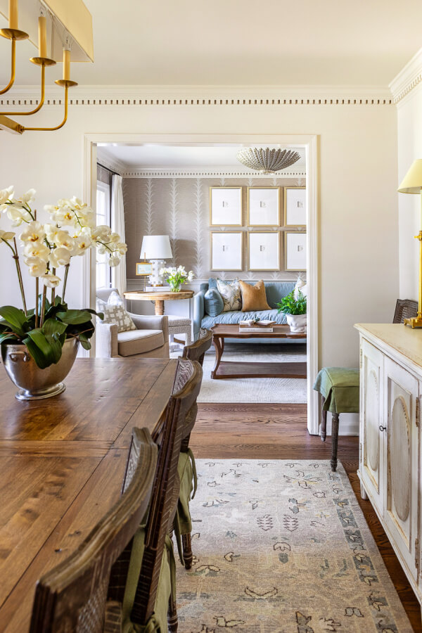 An elegant dining area with a wooden table and chairs leads into a cozy living room with a sofa and framed artwork on the wall. Orchids decorate the table, and there's a chandelier overhead.