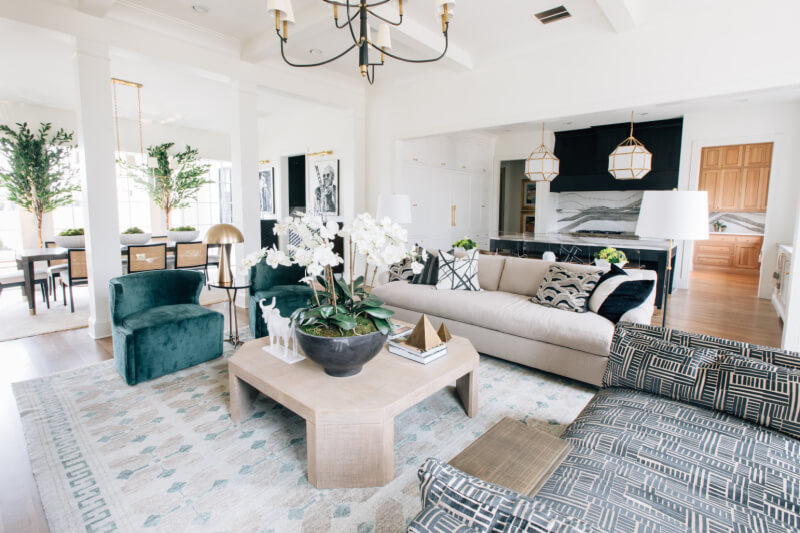 Modern living room with a beige sofa, green armchair, and patterned chair. A wooden coffee table adorned with orchids and a geometric rug enhance the space. The open layout provides views of the dining area and kitchen in the background.