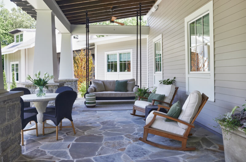 The cozy covered patio showcases stone flooring and features a swing, rocking chairs, a table with black wicker chairs, potted plants, and a charming view of neighboring homes.