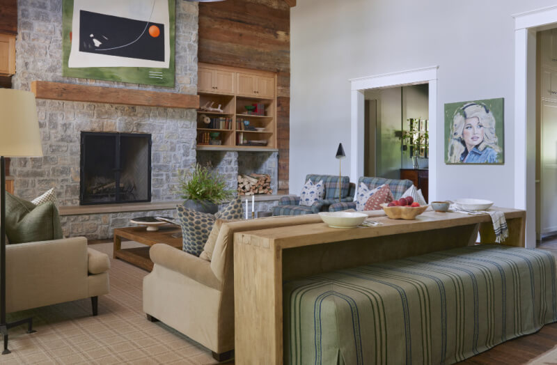 This rustic living room features a stone fireplace, beige sofas, and a wooden coffee table. A large art piece above the mantle complements another painting and bookshelves.