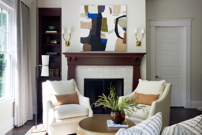 This living room features two white armchairs, a round table with a plant, and abstract art above a dark wood fireplace. Bookshelves and wall sconces flank either side, creating an inviting oasis.