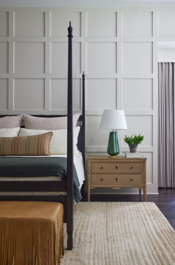 A serene City Hill retreat, this bedroom features a four-poster bed with beige and green bedding, complemented by a wooden nightstand with a lamp and plant. A fringed ottoman adds charm, while the grid-patterned accent wall completes this tranquil oasis.