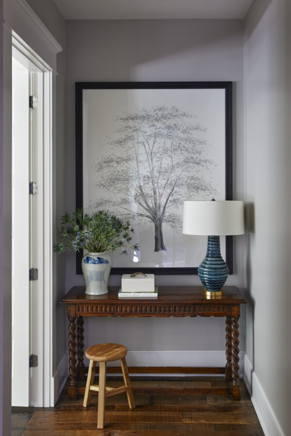 A hallway showcases a wooden table with a stool, a framed tree illustration, a ceramic vase with greenery, and a blue-striped lamp.