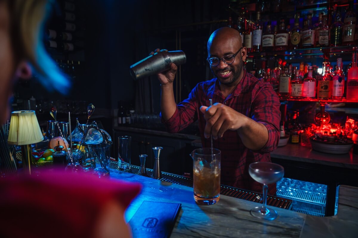 A bartender in a red plaid shirt is smiling as he shakes a cocktail shaker behind a dimly lit bar in Oxford, with various bottles on shelves. This hidden gem is perfect for your Southern City bucket list.