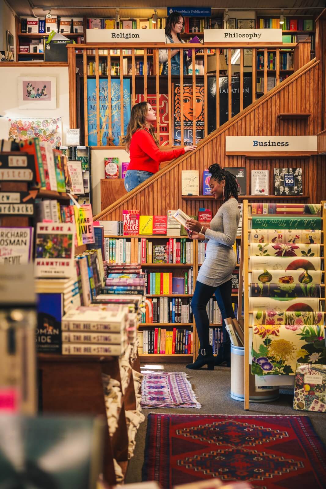 In a cozy Southern city bookstore, two women immerse themselves in literary treasures. One stands on the stairs with a book, while the other is engrossed in reading below, both surrounded by shelves that make this Oxford, MS gem a must-visit on any book lover's bucket list.