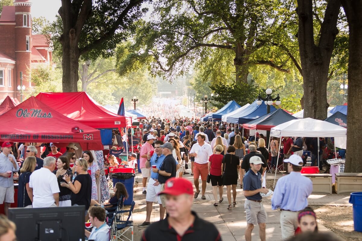 A lively outdoor event in the charming Southern city of Oxford, MS, features numerous people under tents adorned with Ole Miss logos, surrounded by lush trees and a classic brick building. 