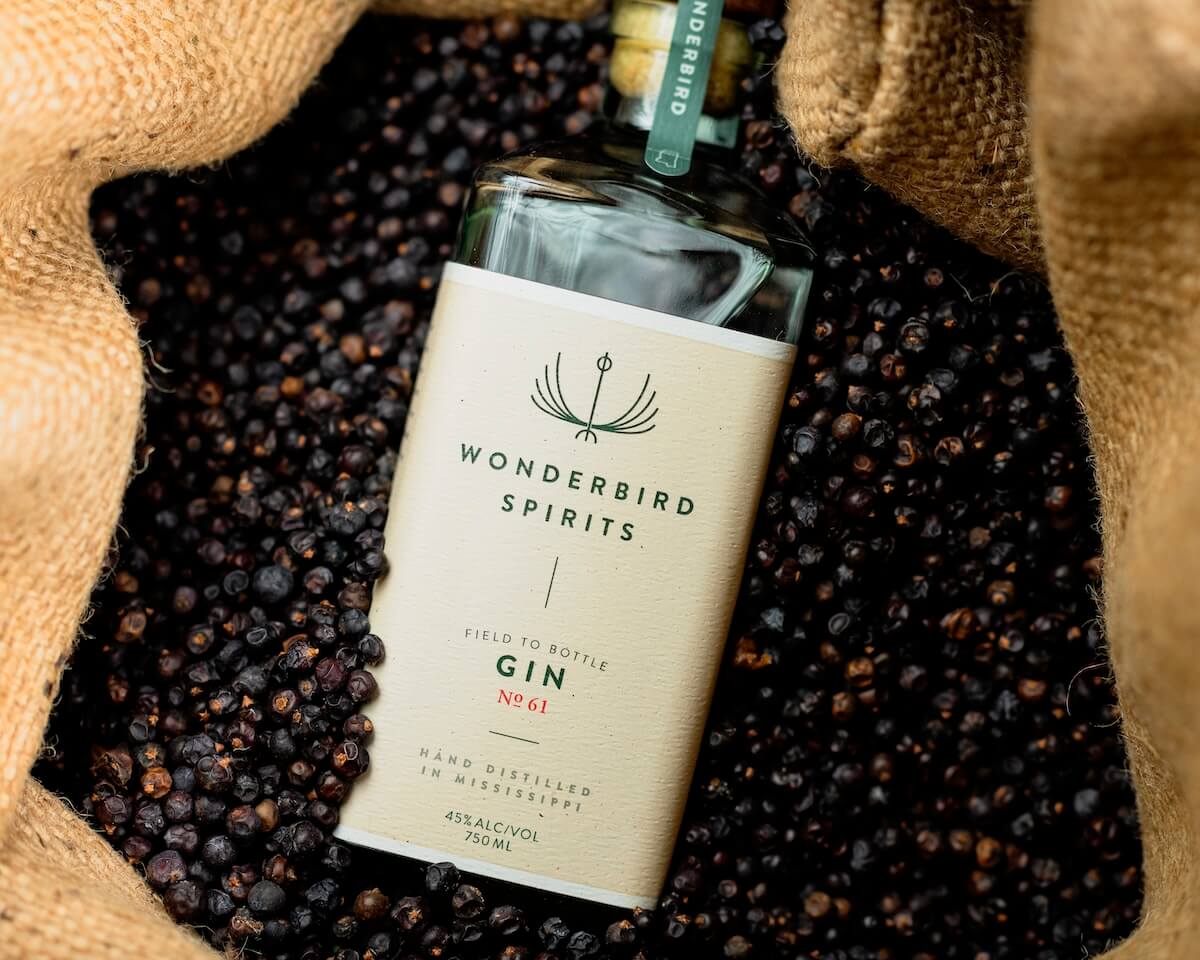 A bottle of Wonderbird Spirits Gin No. 61 rests on a bed of dark berries, partially encased in burlap fabric. Originating from the charming Southern city of Oxford, MS, its label details "45% Alc/Vol, 750 ML.