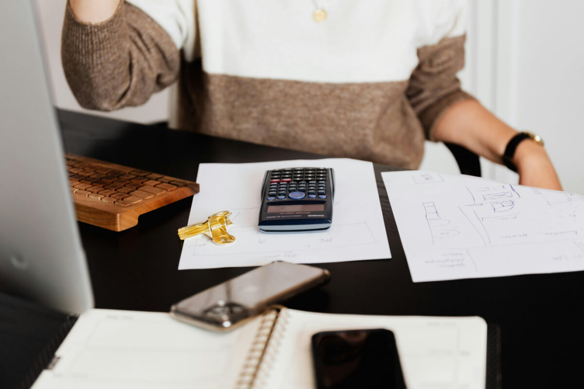 A person sits at a desk amidst a personal finance journey, with a calculator, papers, a phone, a wooden keyboard, and a notebook. Keys rest on one of the sheets of paper, symbolizing the unlocking of financial insights.
