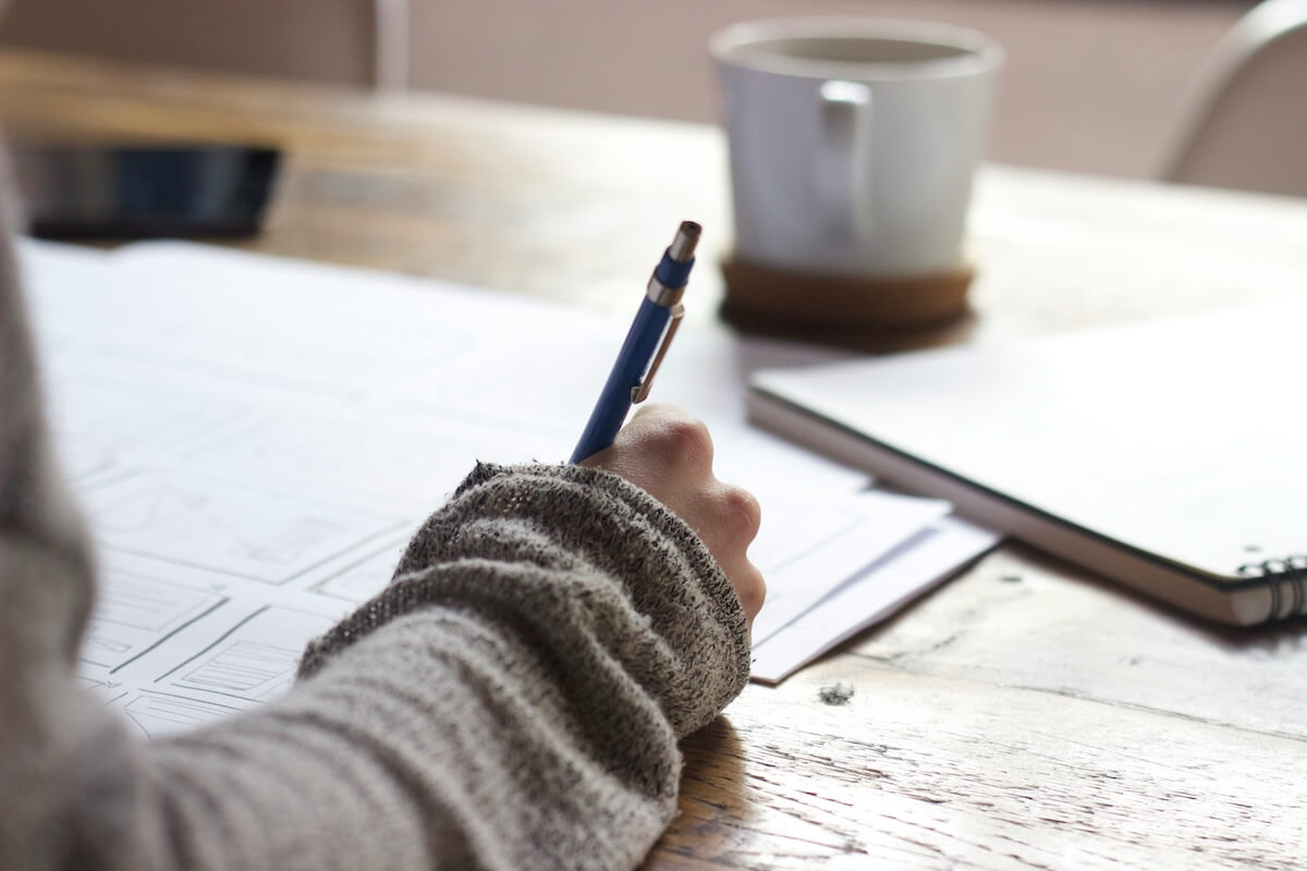A person begins their personal finance journey, writing on paper with a pen, seated at a wooden table. A coffee cup and phone are in the background.
