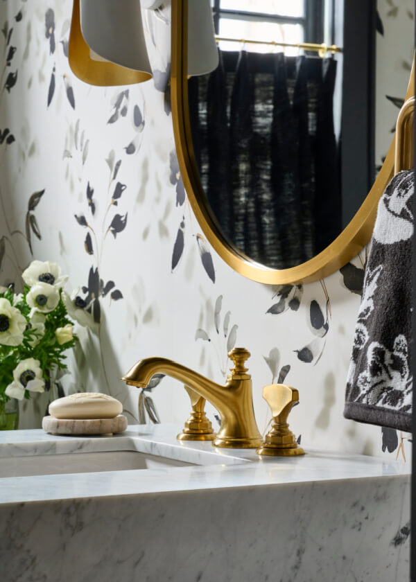 This elegant bathroom sink, a standout home feature, showcases a gold faucet and oval mirror. A marble countertop holds soap and a towel, while floral wallpaper complements a vase with white flowers in the background, creating an exquisite ambiance reminiscent of classic Atlanta charm.