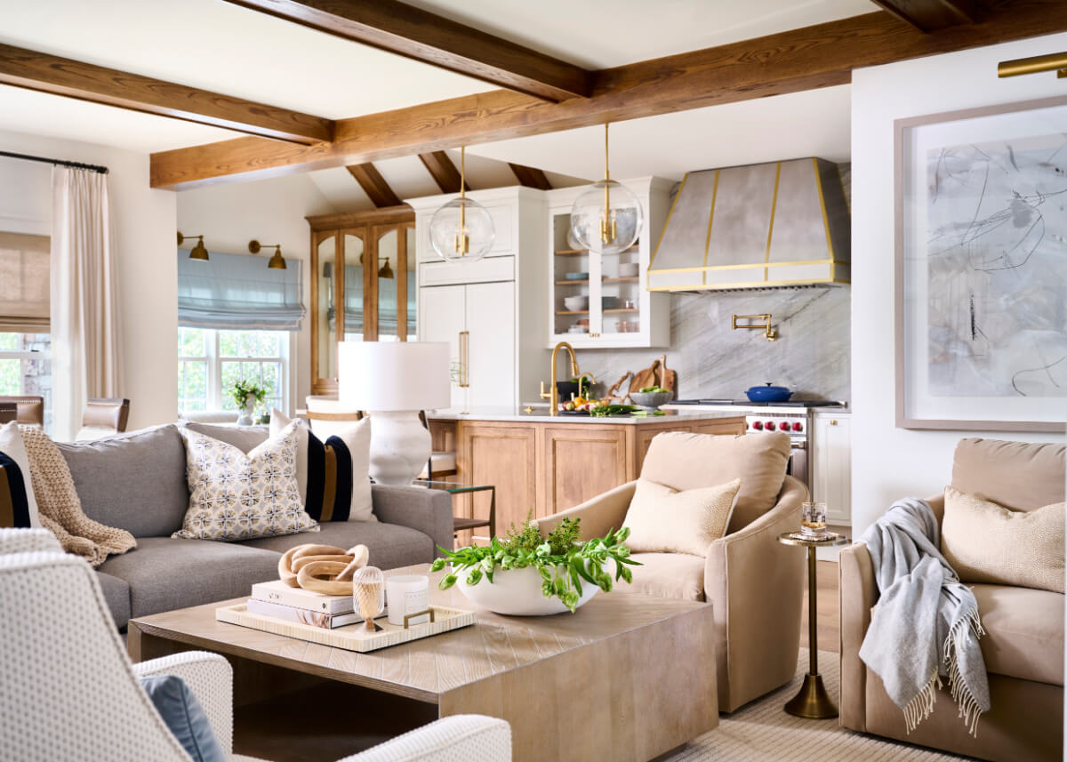 This modern Atlanta home has a living room and kitchen showcasing wooden beams, a gray sofa, beige armchair, and decorative plants. The kitchen's marble backsplash complements abstract art on the wall, while natural light floods in from expansive windows.