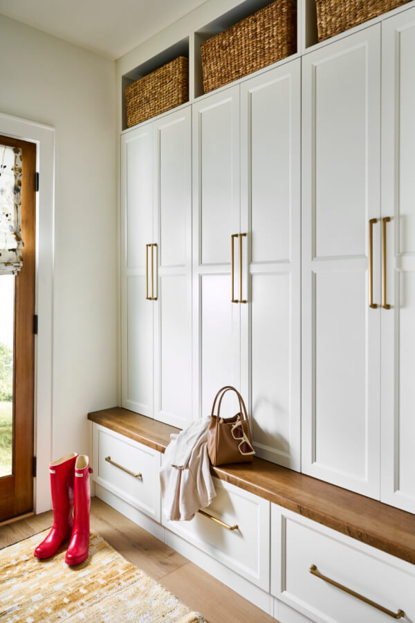 A charming mudroom features white cabinets, a wooden bench, and wicker baskets above. Red rain boots and a beige bag rest by the draped jacket. To the left, a door boasts a patterned curtain, adding to the home's cozy Atlanta charm.