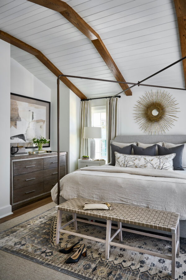 A cozy bedroom in an Atlanta home boasts a canopy bed, patterned rug, and wooden beams on the ceiling. A feature dresser holds a lamp and decor, while a bench and shoes sit at the foot of the bed.