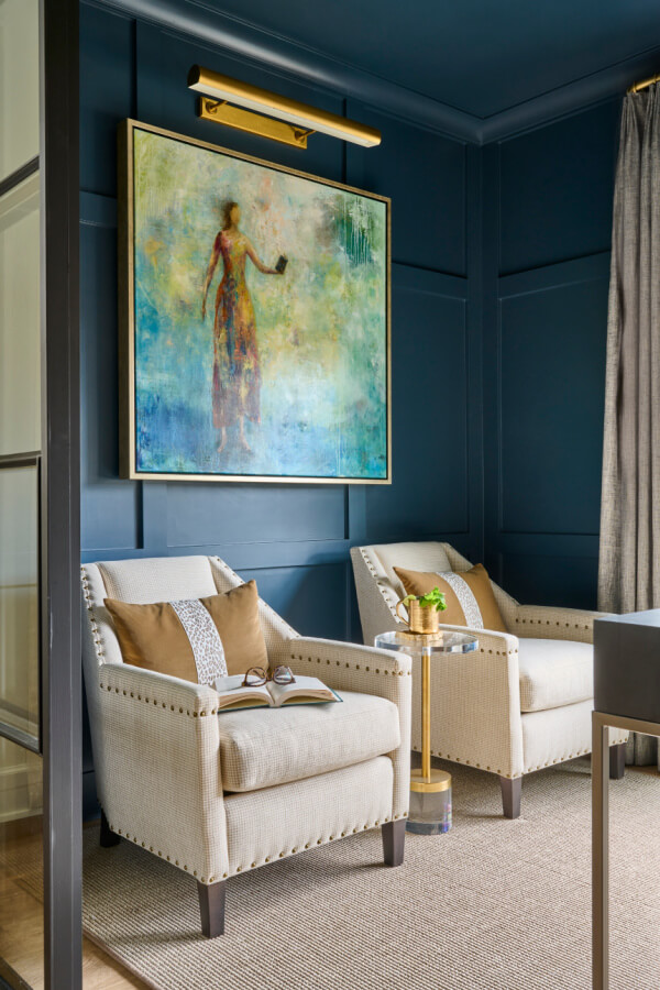 Two beige armchairs with cushions face a side table under a large abstract painting on a dark blue wall, creating a striking feature in this Atlanta home. A window with a curtain is visible on the right.