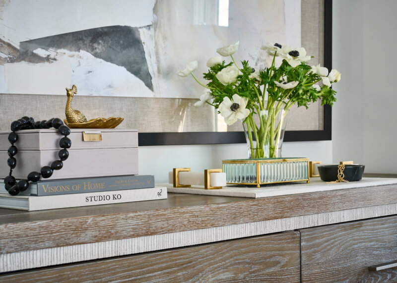 Wooden dresser adorned with books, a beaded necklace, and a decorative box sets the scene in this Atlanta home. A glass vase with white flowers and a tray with a metal handle add charm, while mounted abstract artwork completes this captivating home feature.