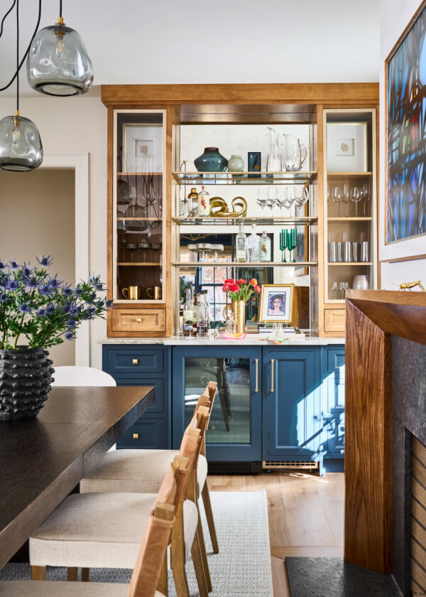 In this Atlanta home, the dining room showcases a blue and wood hutch with glassware, a small fridge, and decor. A dark wooden table with light chairs sits nearby. Hanging pendant lights and a stained-glass window feature prominently, enhancing the ambiance.