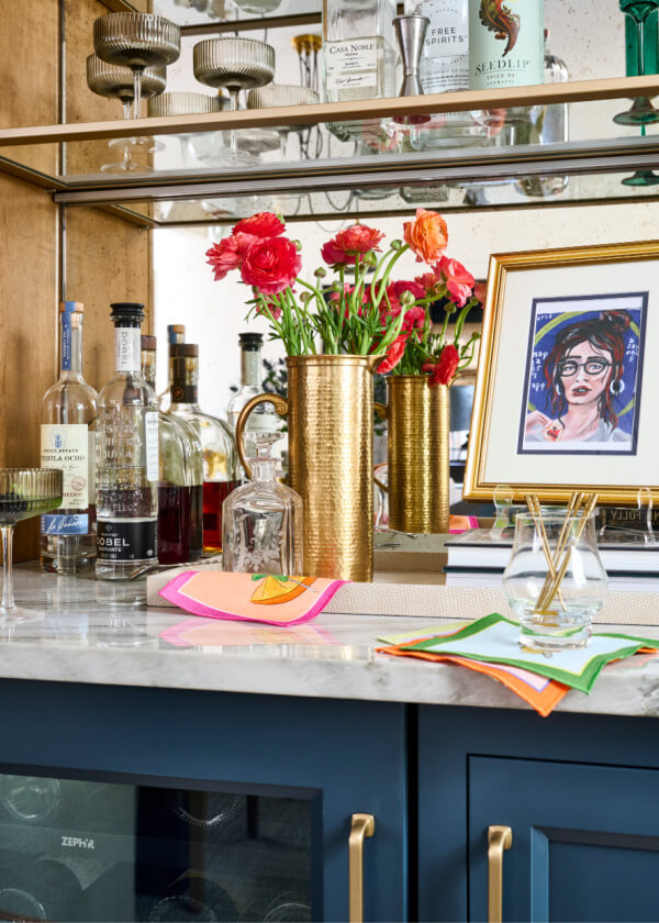Experience the Atlanta charm in this exquisite home bar setup, featuring a marble counter, an impressive display of various liquor bottles, a golden vase with red flowers, and a mirrored backsplash. A framed portrait adds a sophisticated feature to this elegant space.