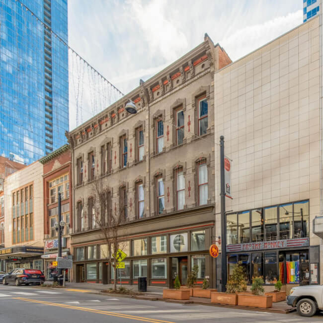 Street view in Nashville with a mix of historic and modern buildings, featuring a three-story old structure with visible wear and a contemporary glass high-rise in the background. Sidewalk planters add charm to these iconic Nashville landmarks.