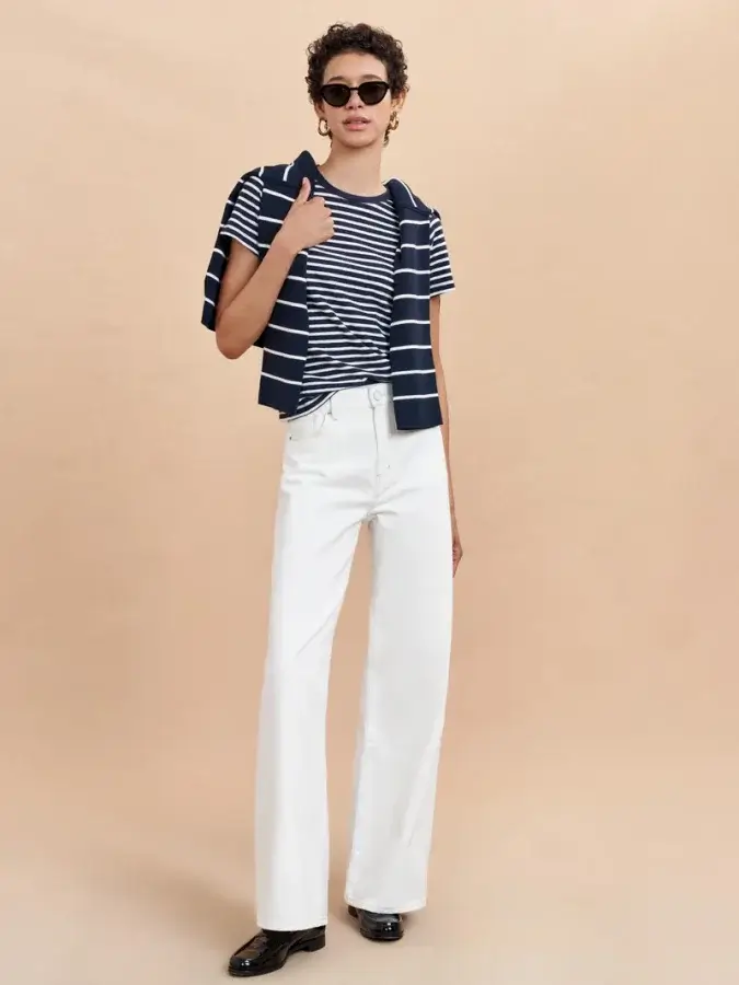 A person sporting sunglasses and white pants stands confidently against a neutral background, exemplifying modern spin with a striped t-shirt and matching sweater draped over the shoulders—a true nod to spring wardrobe essentials.