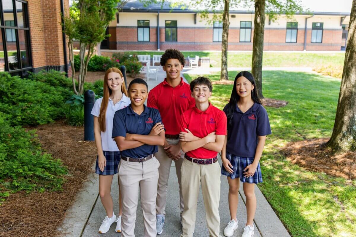 Nashville Private School Open House Dates - 3 Five students stand outdoors at a school campus, wearing matching uniforms with navy or red shirts and plaid skirts or khaki pants, smiling at the camera.
