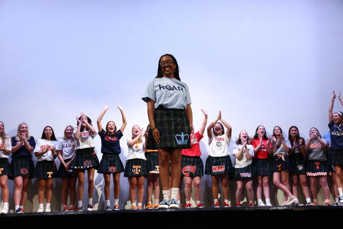 Nashville Private School Open House Dates - 13 A group of young women in skirts and college t-shirts stand on stage, with one woman in front wearing a "ROAR" shirt while others clap and cheer behind her.