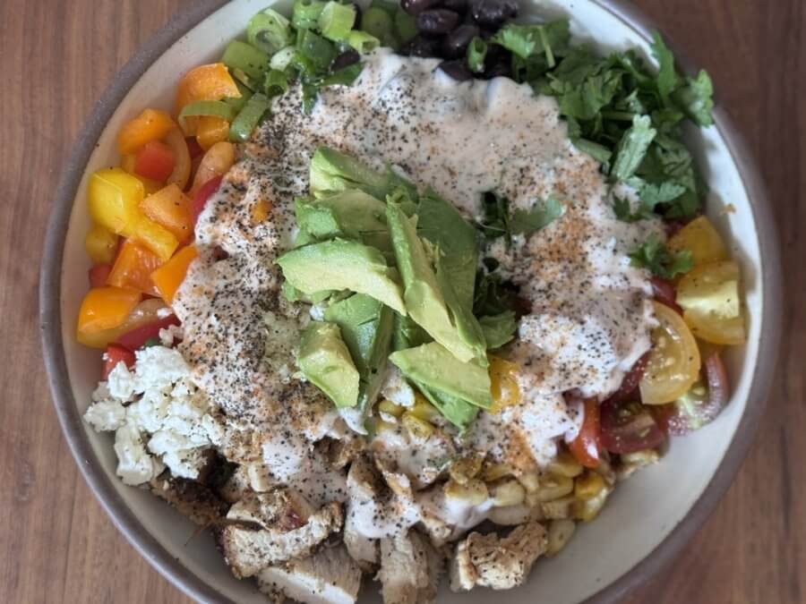 A bowl brimming with diced vegetables, black beans, grilled chicken, chopped greens, and avocado slices drizzled with creamy dressing—a perfect choice for those seeking high protein recipes.